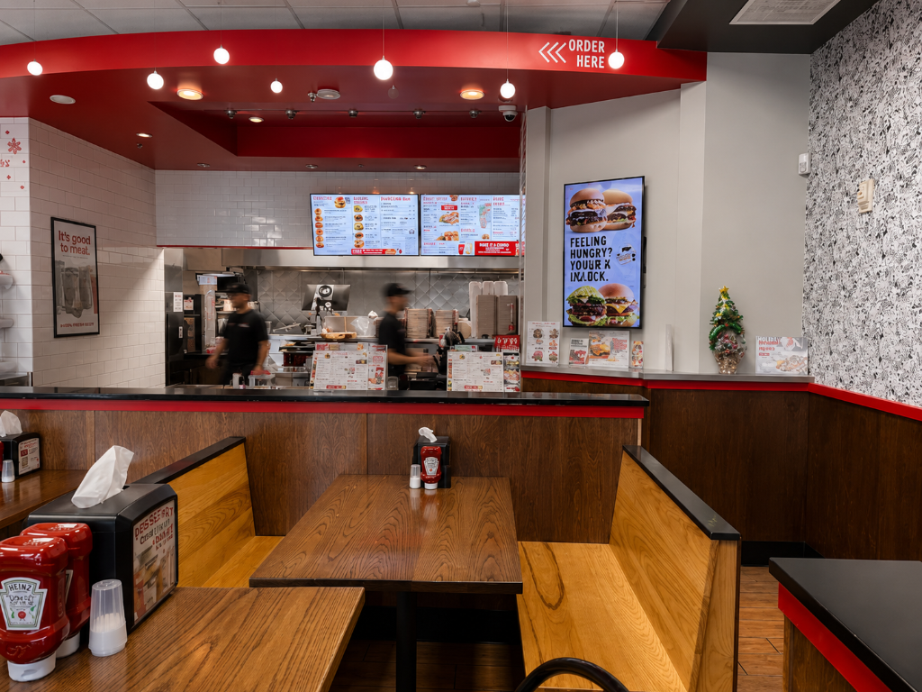 Interior of Mooyah burger franchise in Winter Park, FL, showing a long dining aisle with white brick walls, red metal chairs, wood-topped tables, built-in booth seating, and a dark charcoal ceiling soffit with red pendant lights