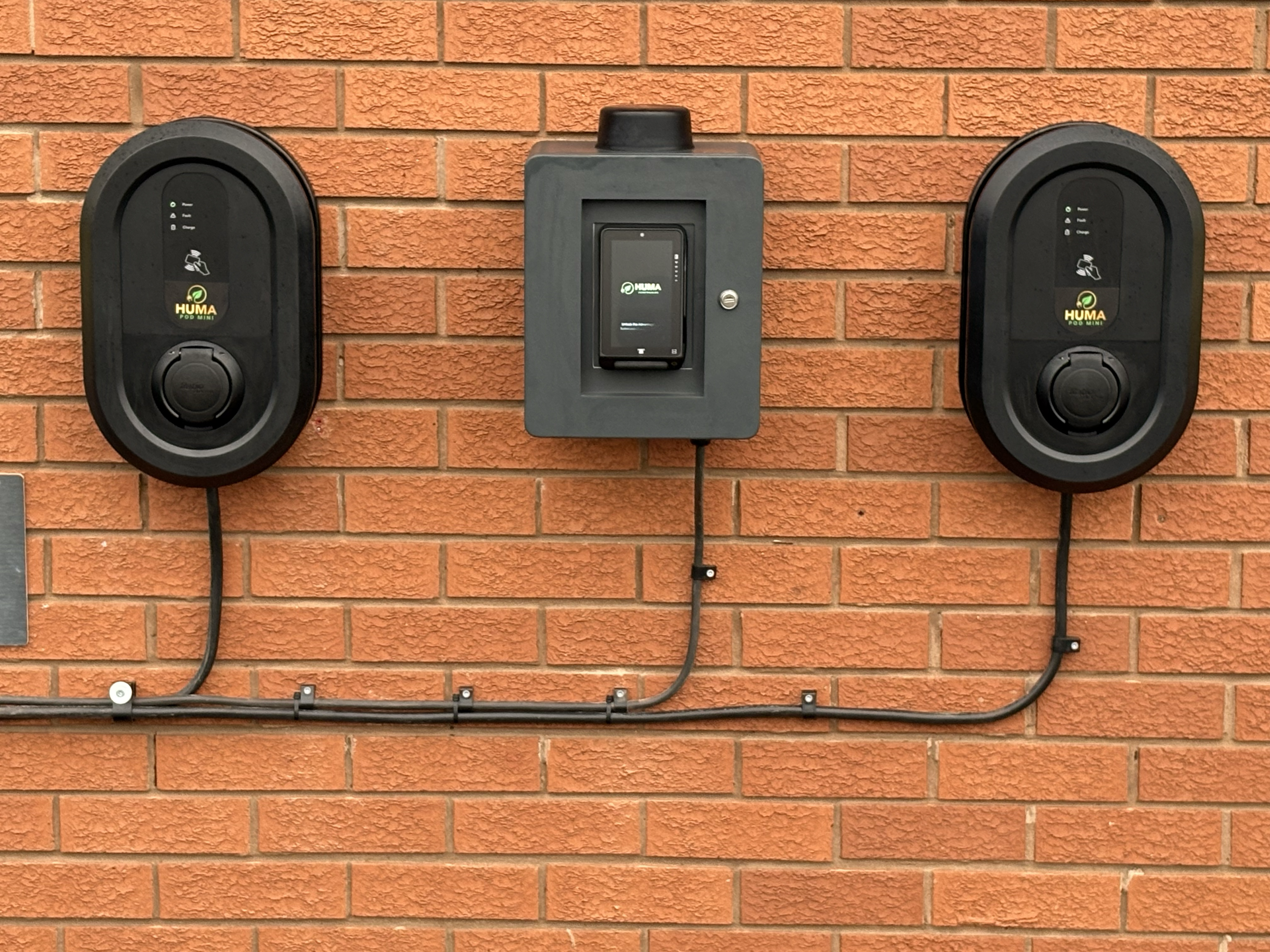 A professional installation of two HUMA Pod Mini electric vehicle chargers flanking a central HUMA control terminal on a red brick wall