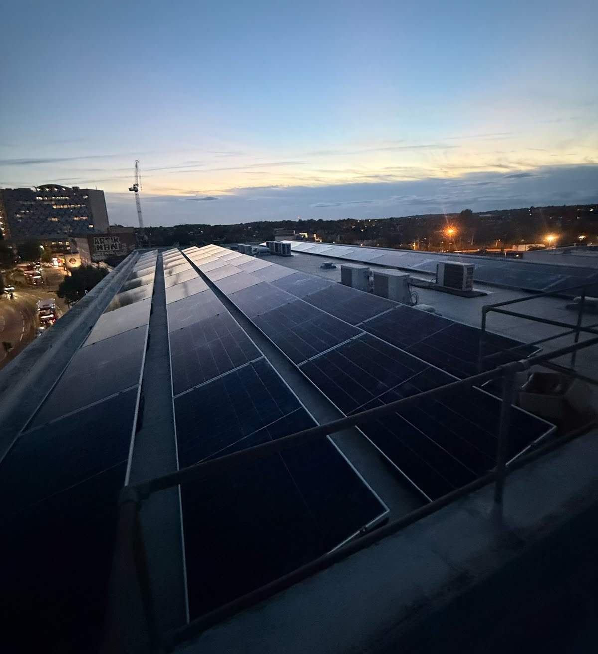 Elevated view at dusk of a rooftop solar PV array with city buildings and a construction crane in the background