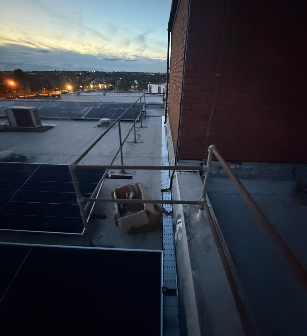 High-angle view across a flat rooftop at dusk, with rows of solar panels and a brick wall and safety railing in the foreground