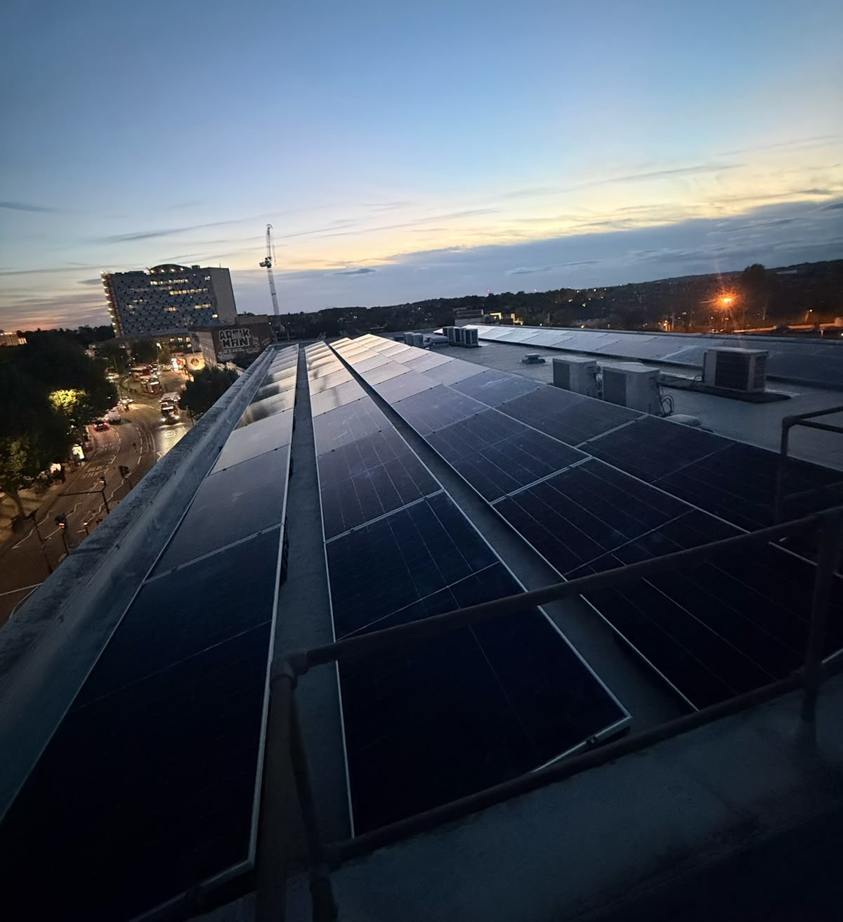 High-angle view of a flat-roof solar PV array at twilight, with parallel rows of panels and an urban skyline in the distance
