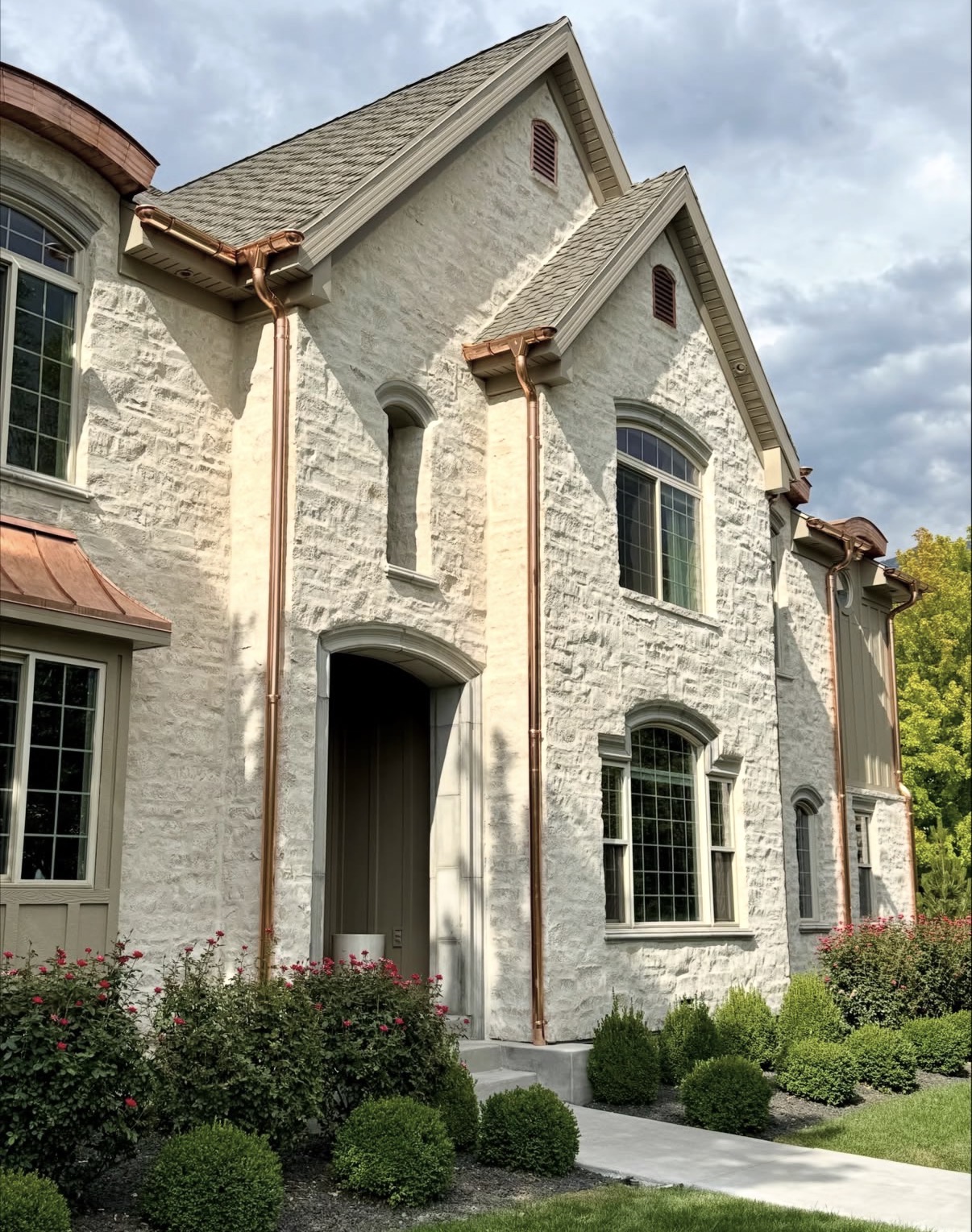 Front view of cream stone masonry home with brown-grey asphalt shingle roof and polished copper gutters and downspouts
