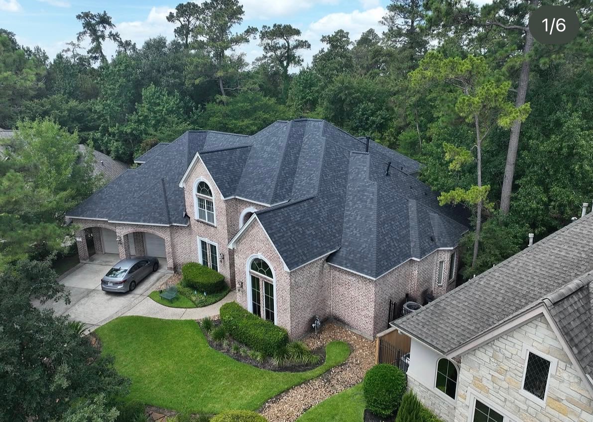 Drone view of large two-story home with dark charcoal architectural shingle roof and tan brick facade