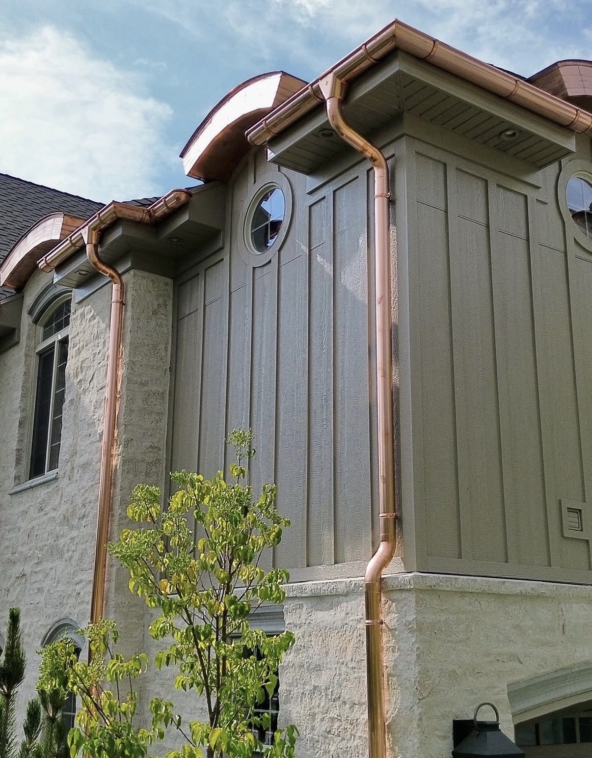 Low-angle view of multi-story home with olive-taupe board-and-batten siding, stone foundation, and polished copper gutters and downspouts