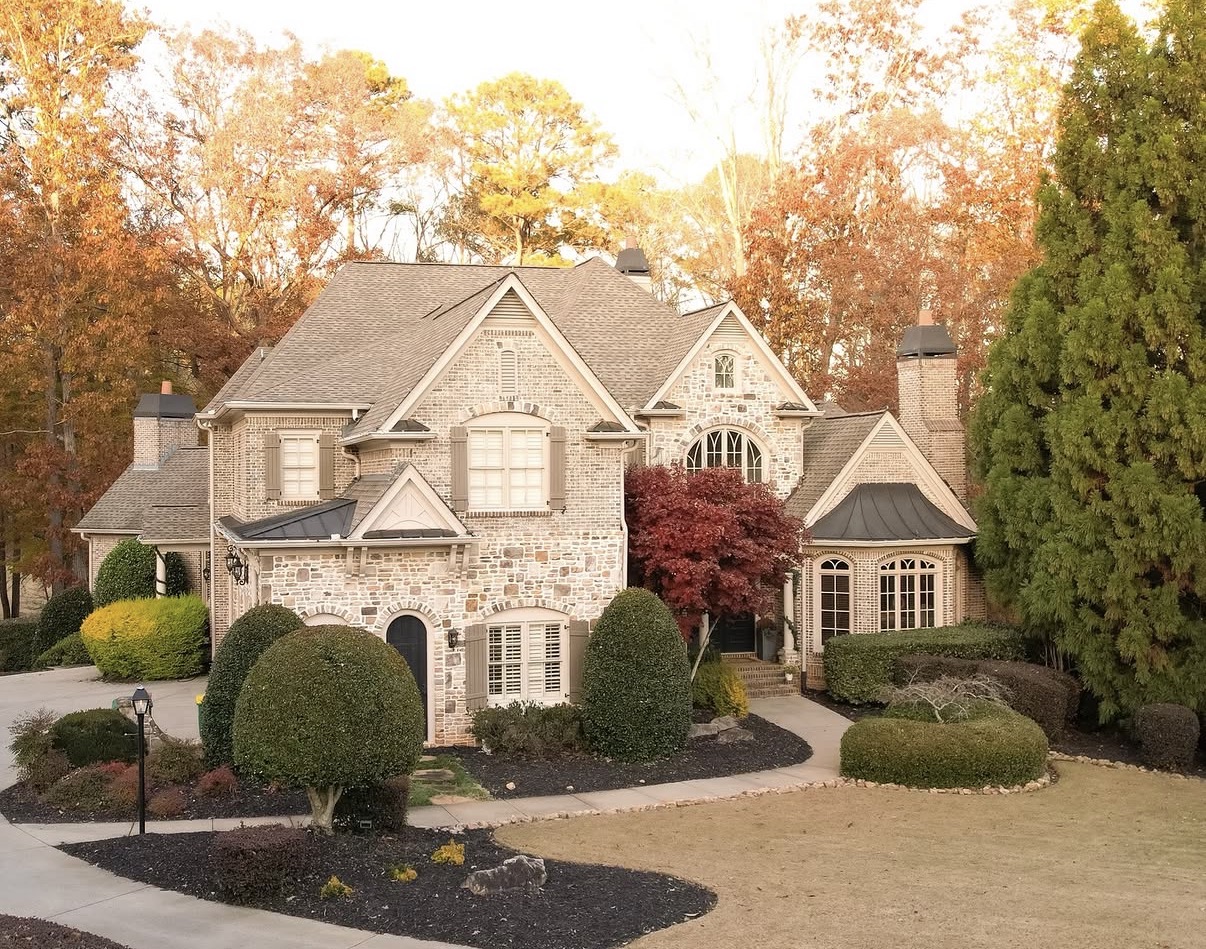 Elevated view of large two-story brick and stone home with brown-grey asphalt shingle roof and charcoal metal roofing accents
