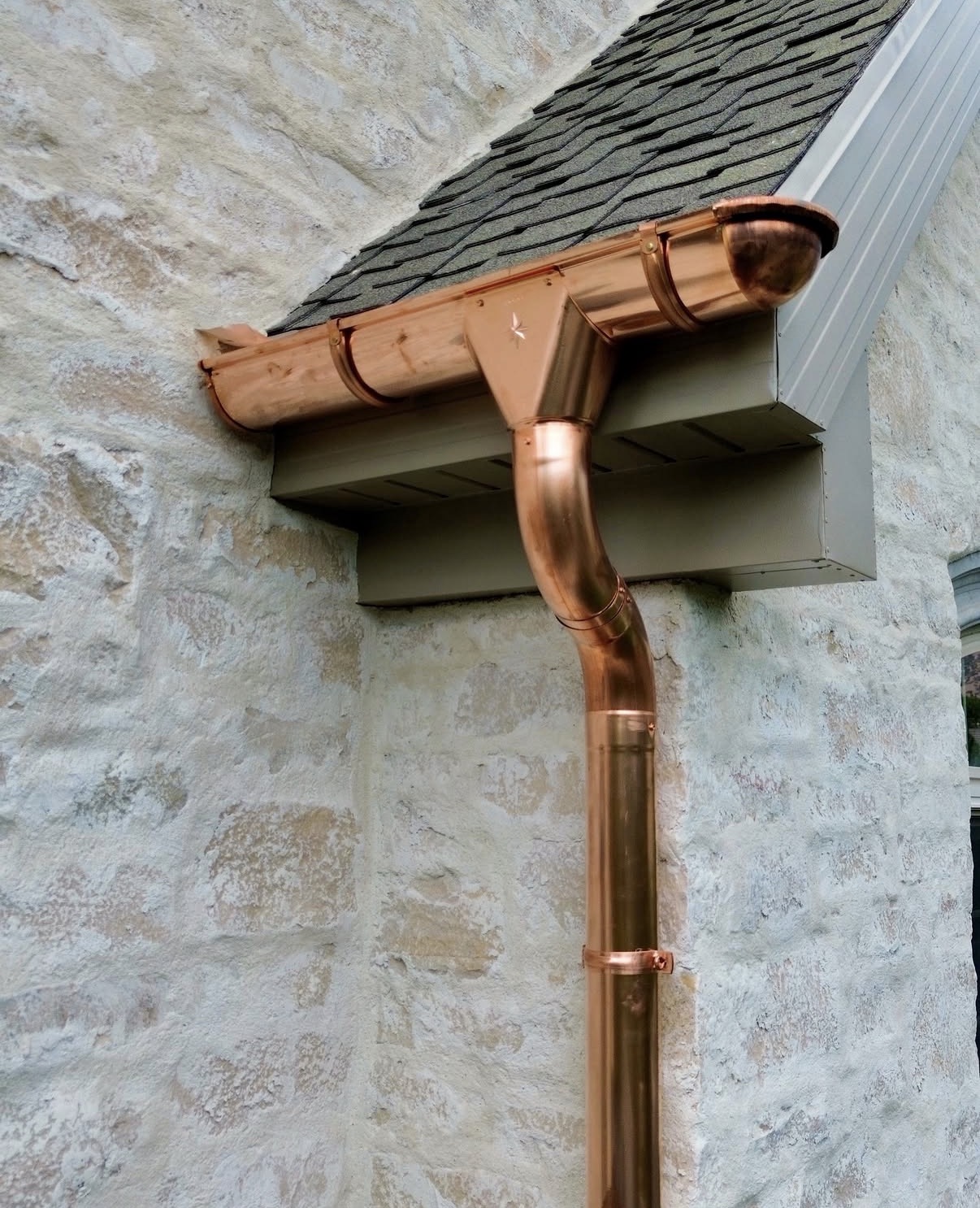 Close-up of newly installed polished copper K-style gutter and downspout with elbow fitting against a cream stone wall