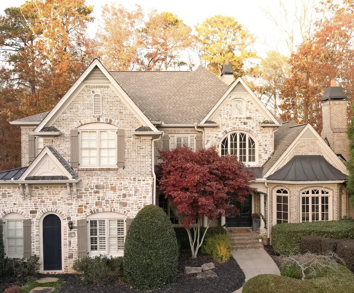 Two-story stone and brick home with charcoal-gray architectural shingle roof and dark bronze metal roof accents over entryway