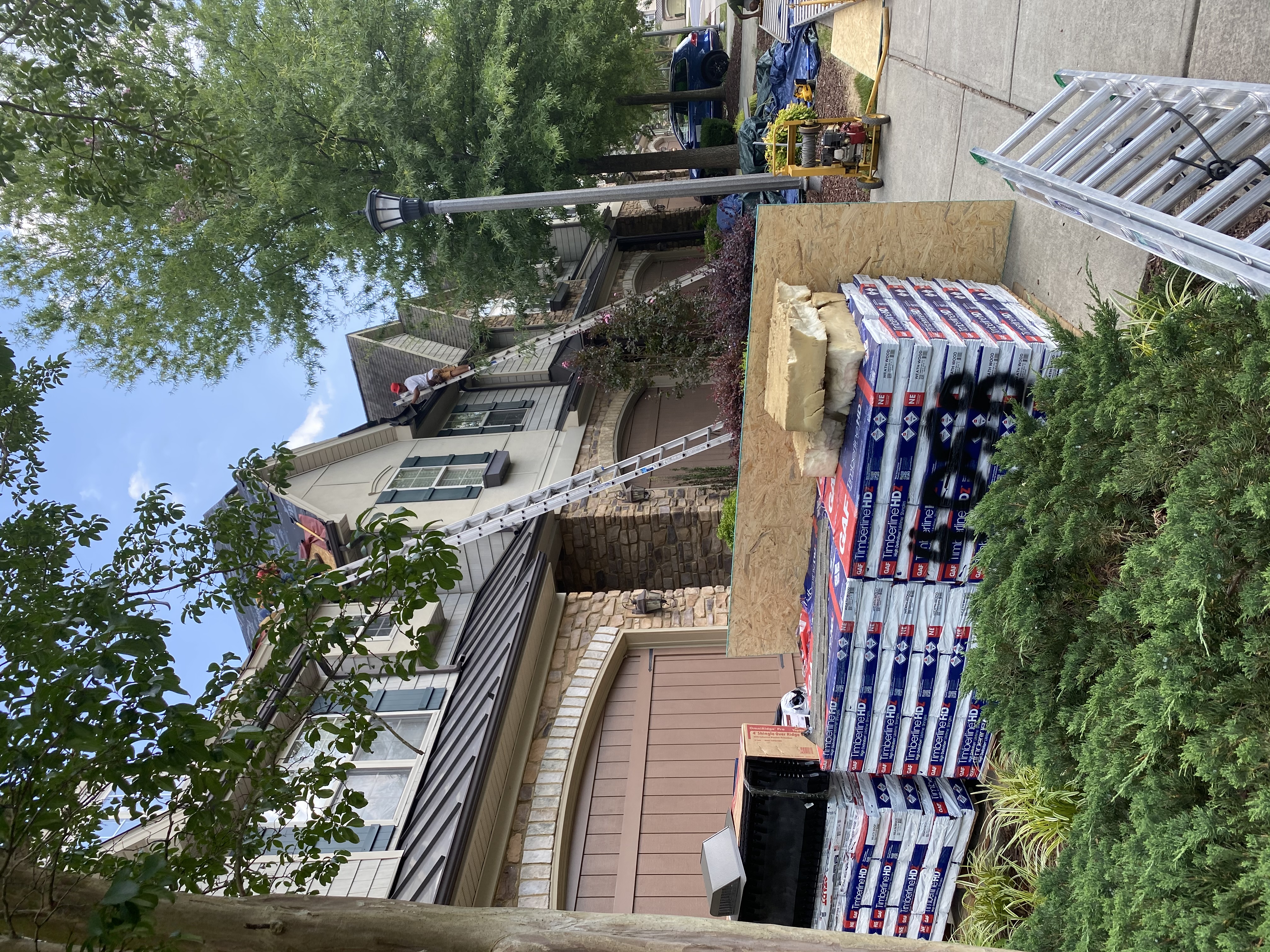 Roofing project in progress on stone veneer home with GAF Timberline HD shingle bundles staged and crew working on the roof