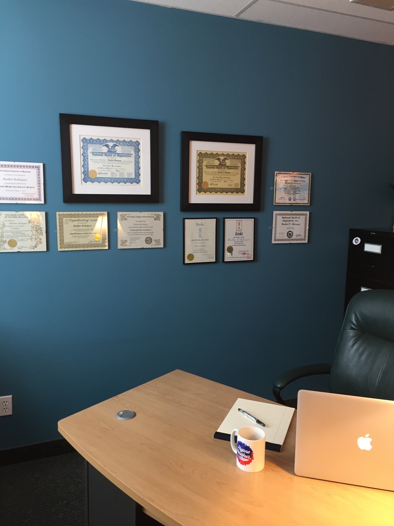 Heather's Regina office desk with framed certifications, laptop, and Hypno Heather mug