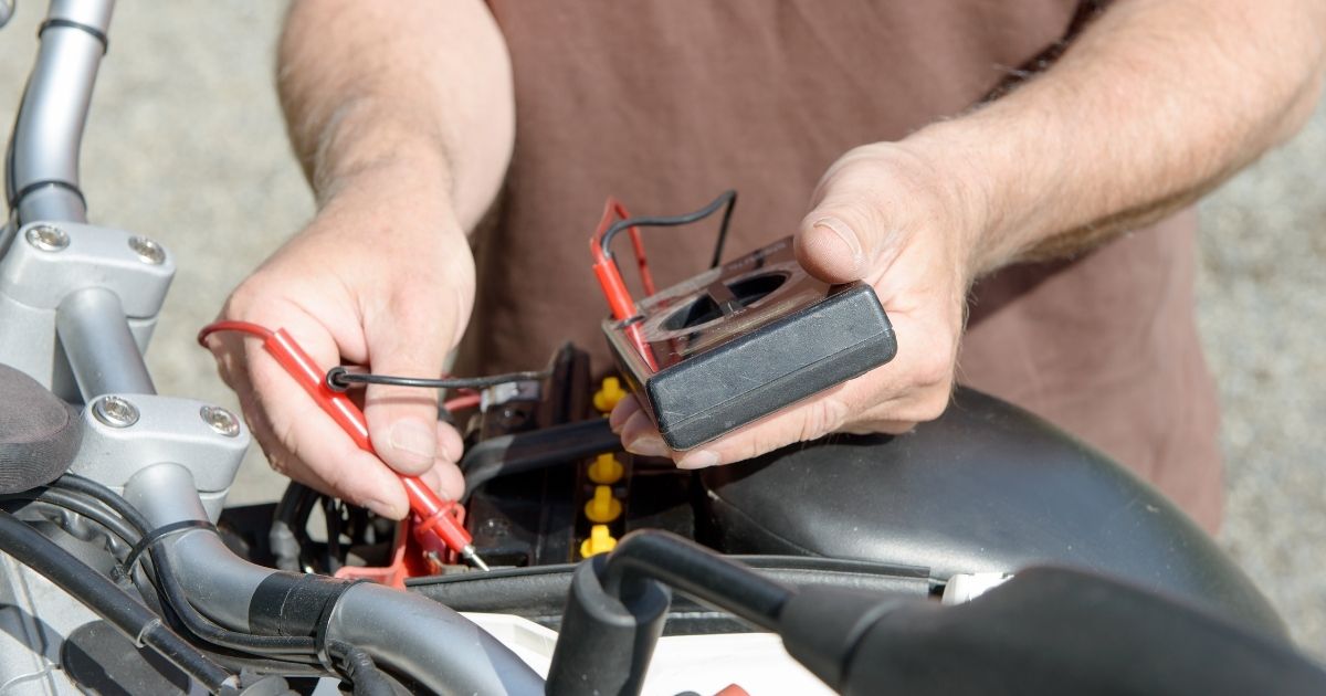 Tangan menguji voltan bateri motosikal menggunakan multimeter digital di atas hendal motosikal