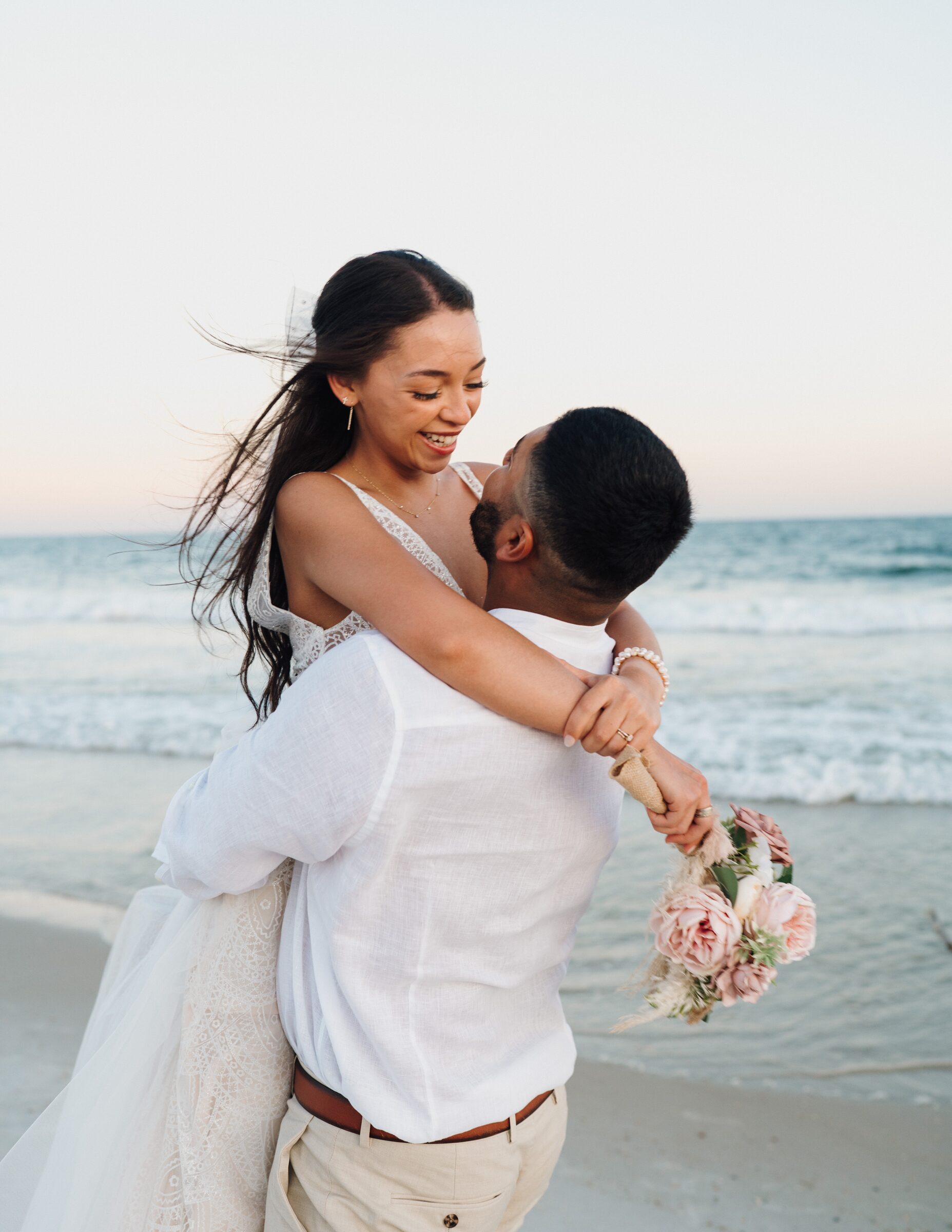 Groom lifting the bride in an embrace on the beach at a sunset wedding ceremony on the Gulf Coast