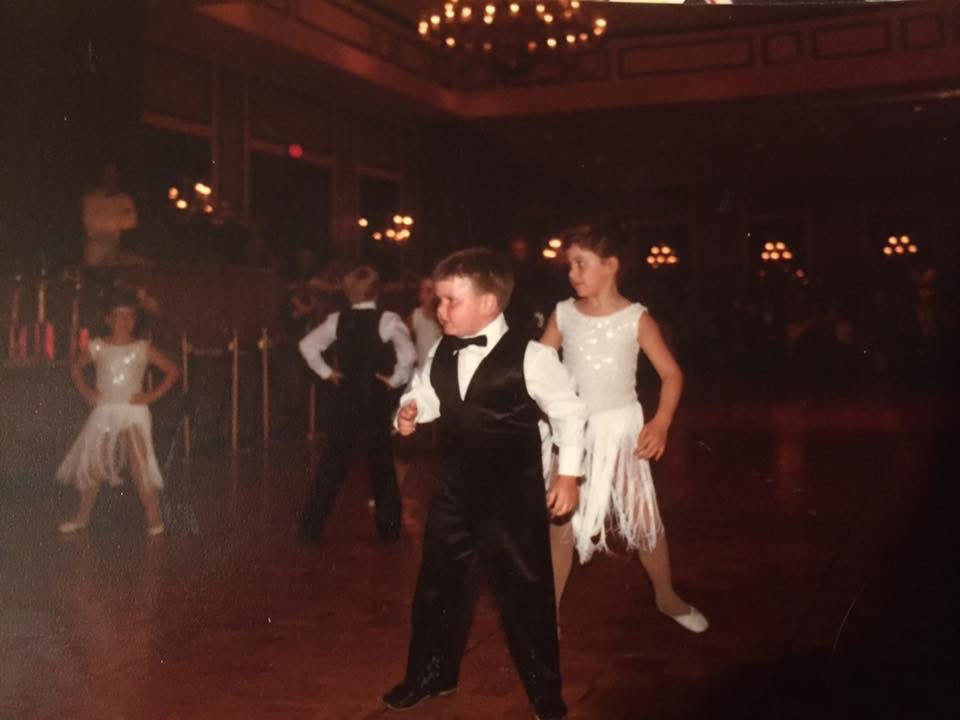 A boy in a black vest and bow tie dancing in a ballroom under chandeliers
