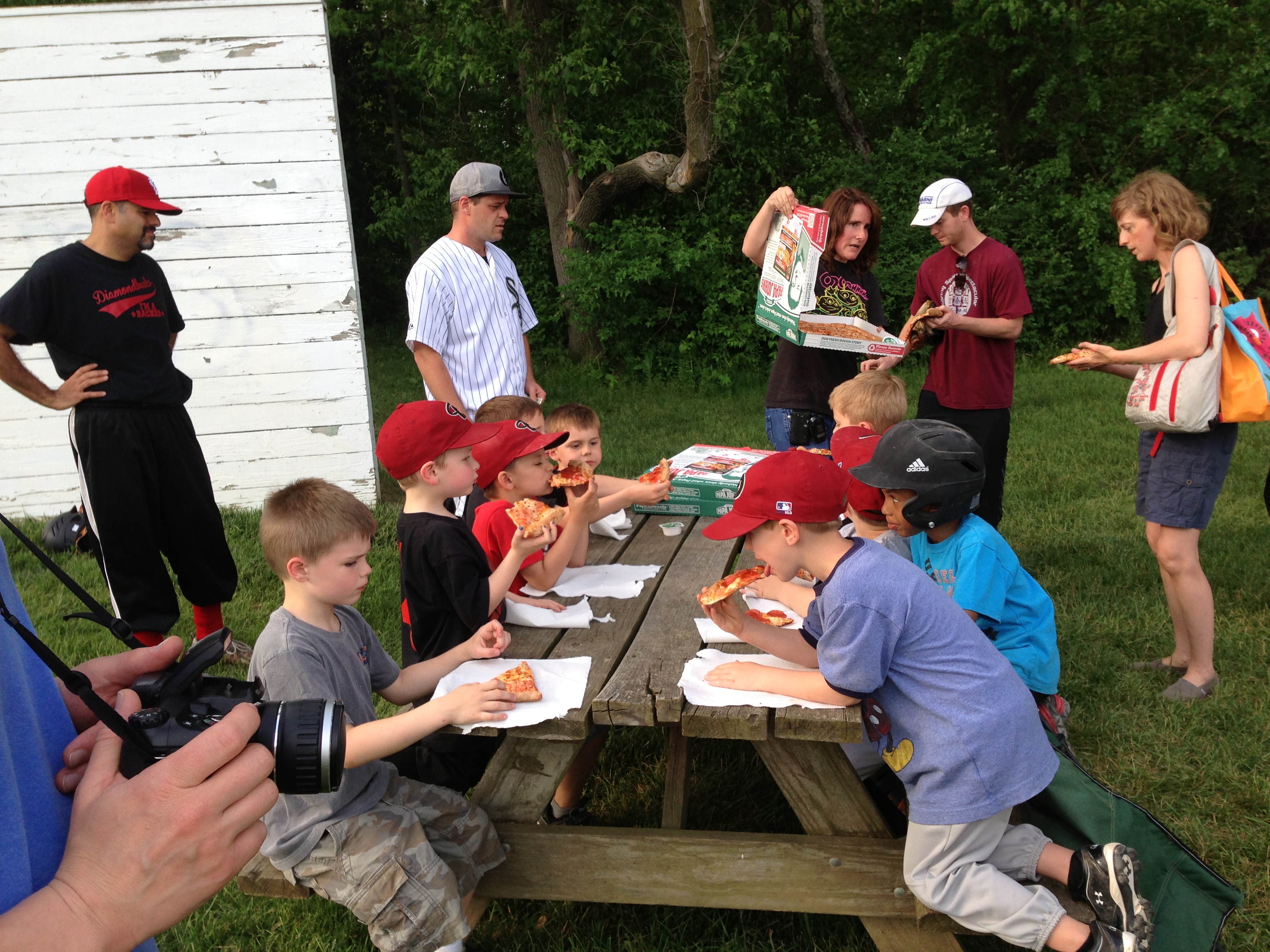 Children eating pizza at a picnic table after a ball game while parents stand nearby.