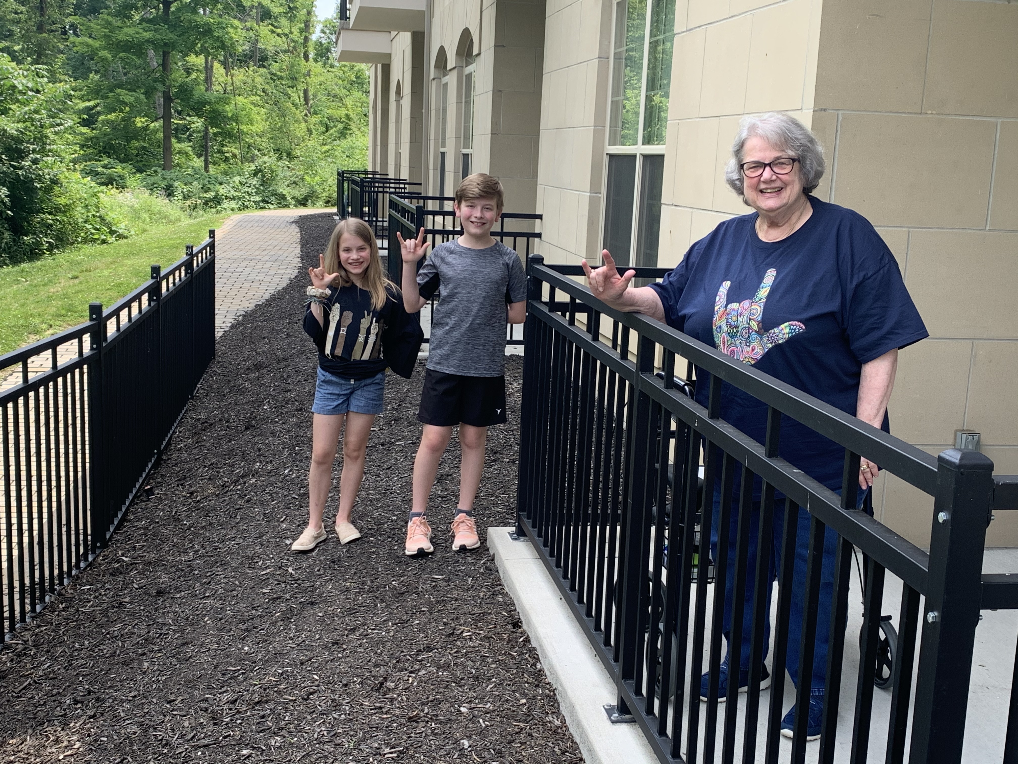 A grandmother and two grandchildren signing "I love you" on a walkway outside a senior residence.
