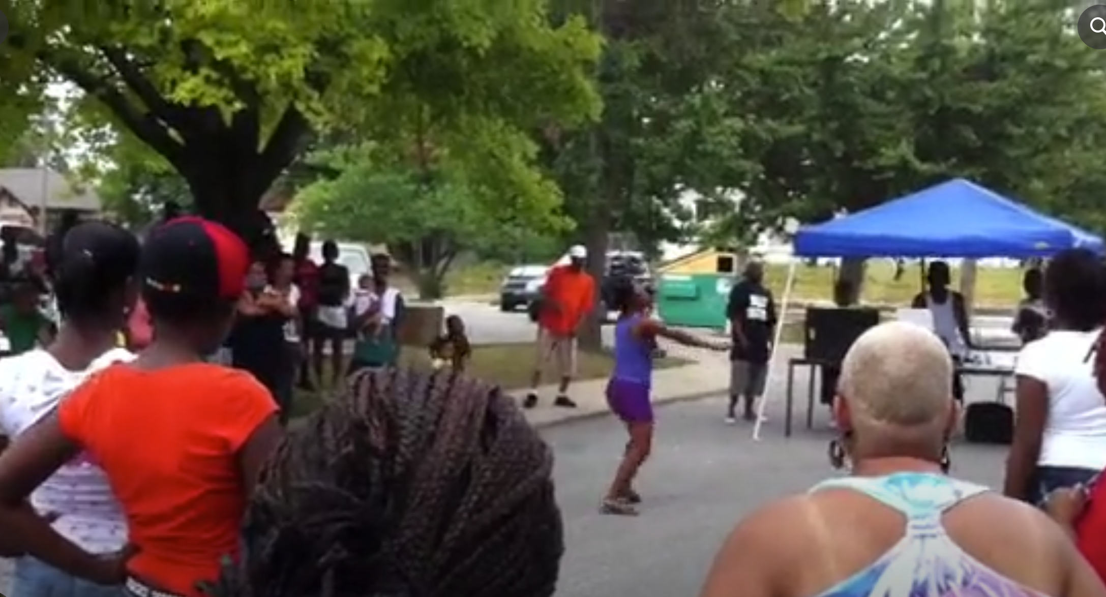 A block party in the street, neighbors watching a girl dance under a canopy tent.