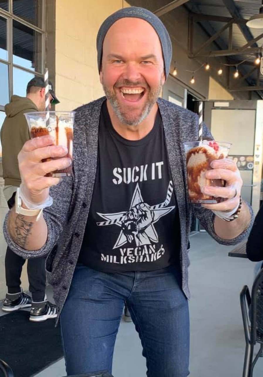 Marc McAleavey grinning, holding up two milkshakes, in a beanie and a "Suck It Vegan Milkshakes" shirt.
