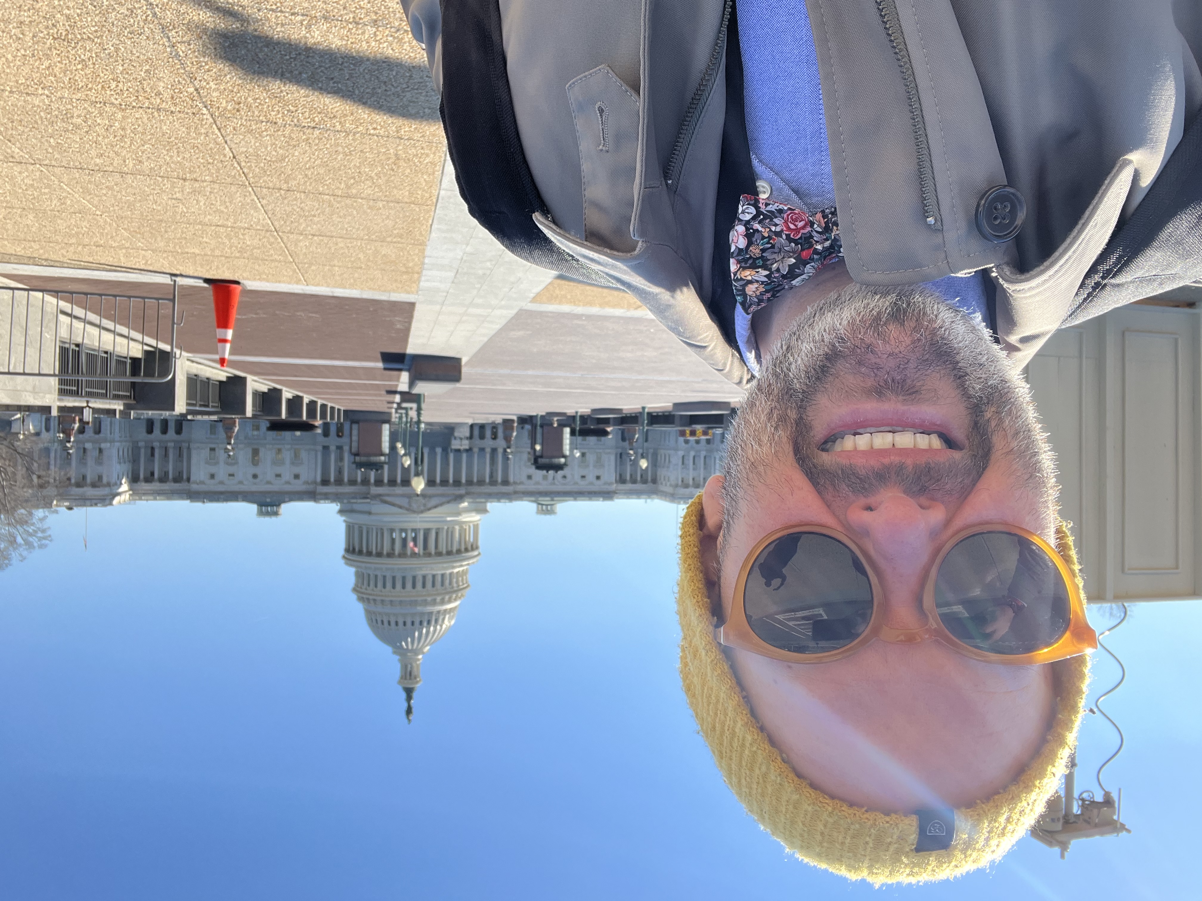 Marc McAleavey smiling in a yellow beanie and sunglasses in front of the United States Capitol on a clear day.