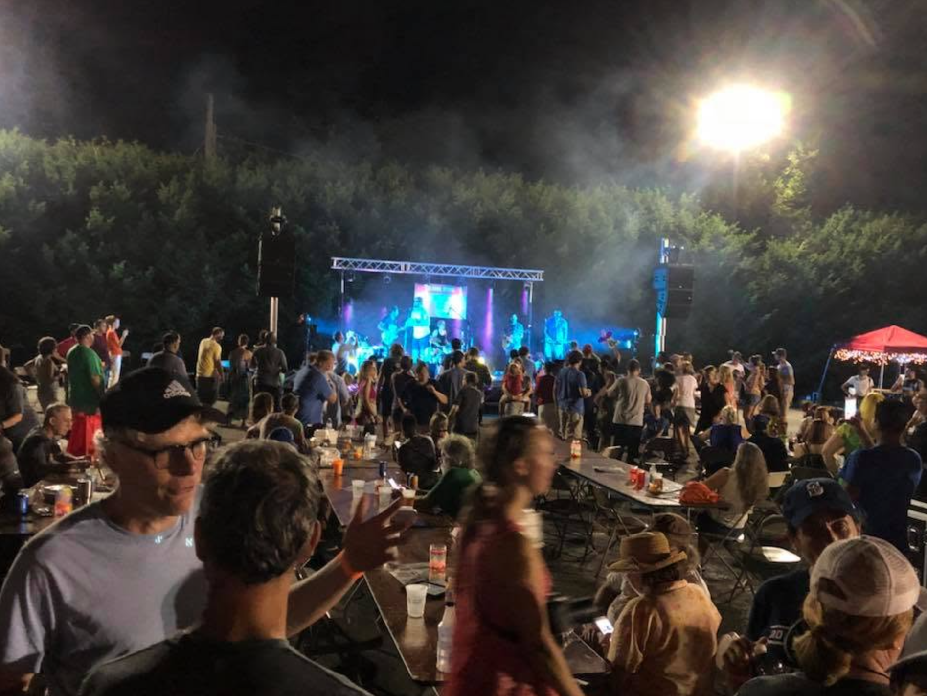 An outdoor concert at night, a crowd at folding tables in front of a lit stage.