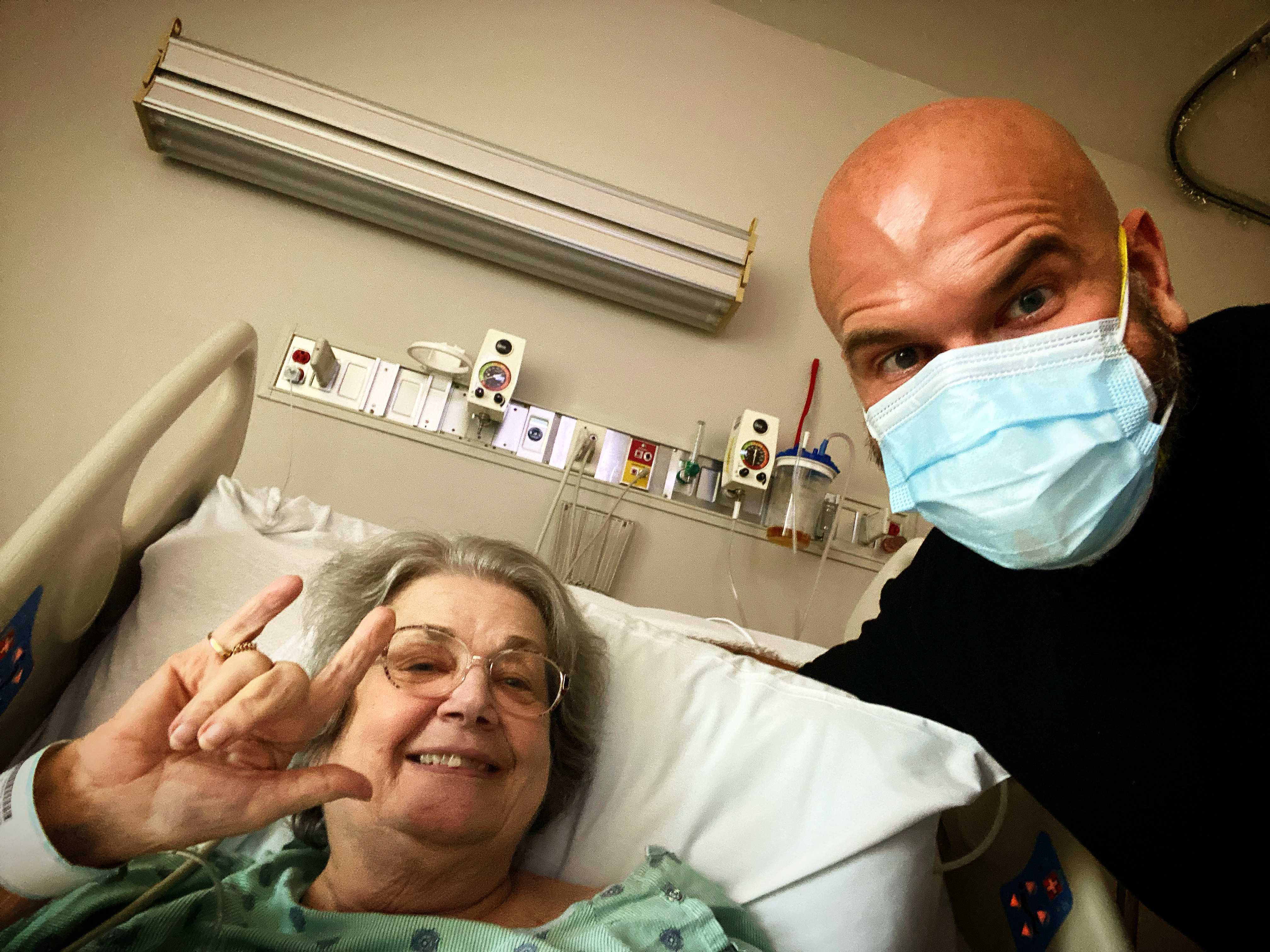 A grandmother in a hospital bed signing "I love you" beside her masked grandson.