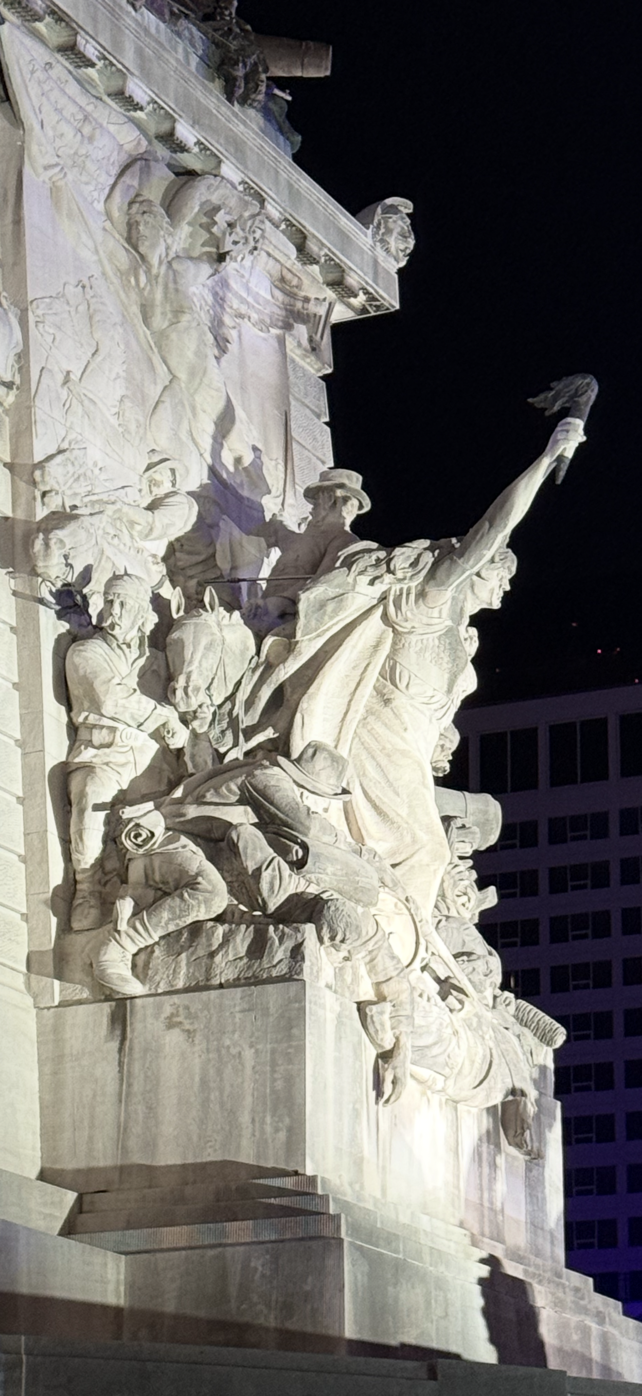 The Soldiers and Sailors Monument in Indianapolis at night: a limestone figure thrusts a torch into the black sky while carved soldiers fall across the stone beneath her.