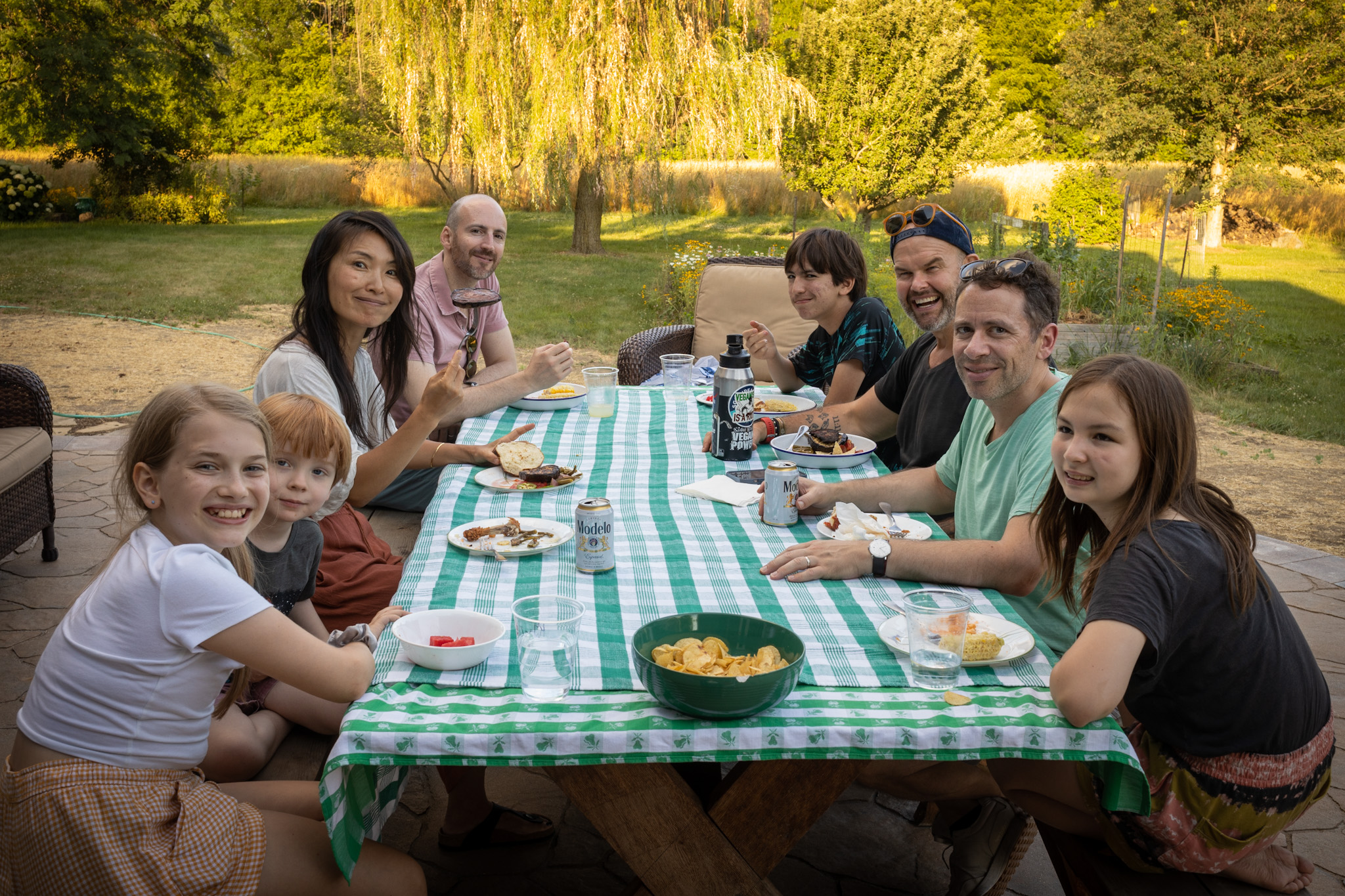 Neighbors of several households sharing a meal at one long green striped table in a backyard.