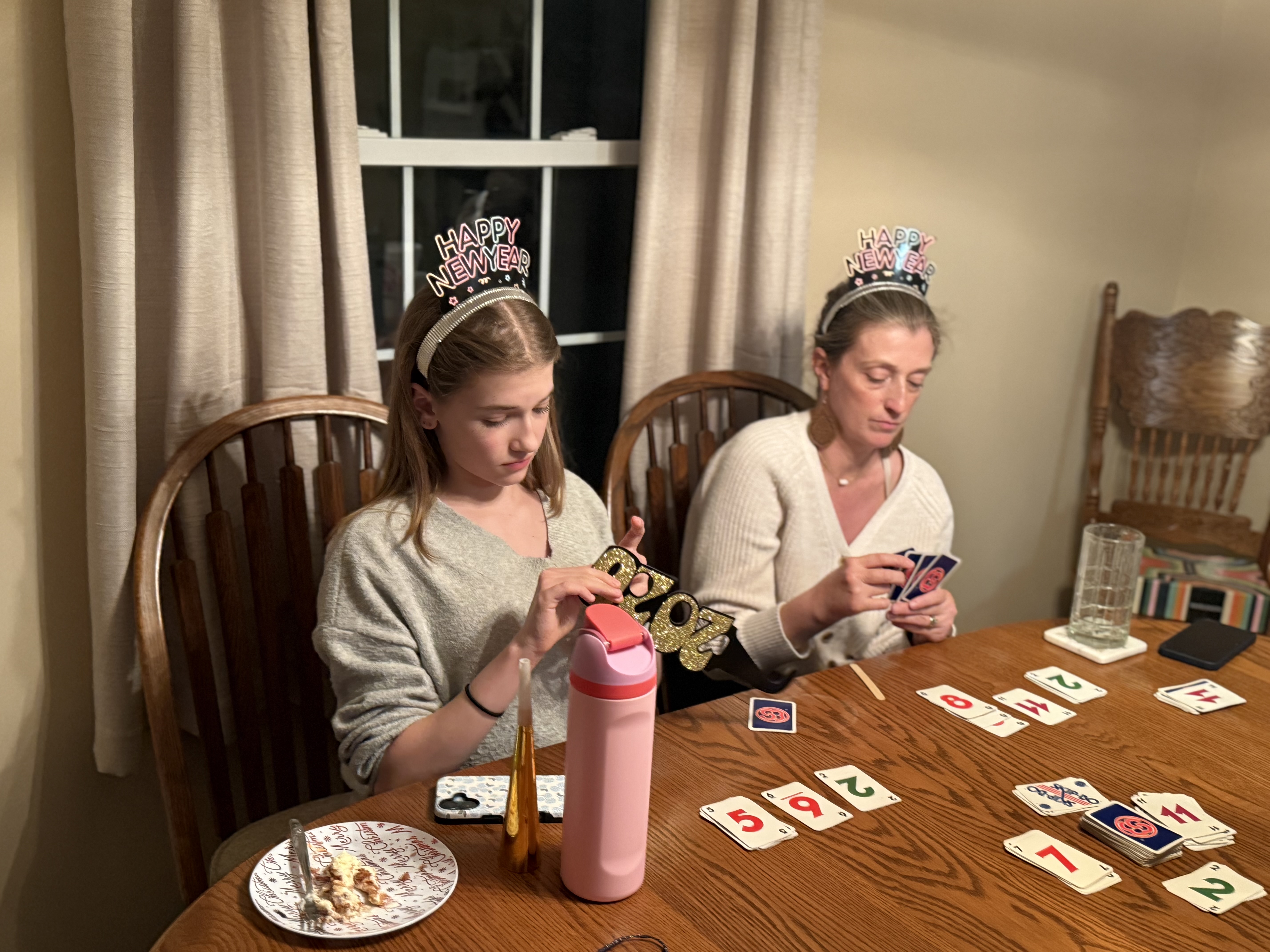 A mother and daughter in Happy New Year headbands playing cards at a kitchen table
