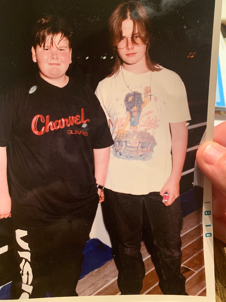 Two boys on a ship deck at night, one in a Charvel Guitars shirt, one in a Mötley Crüe shirt