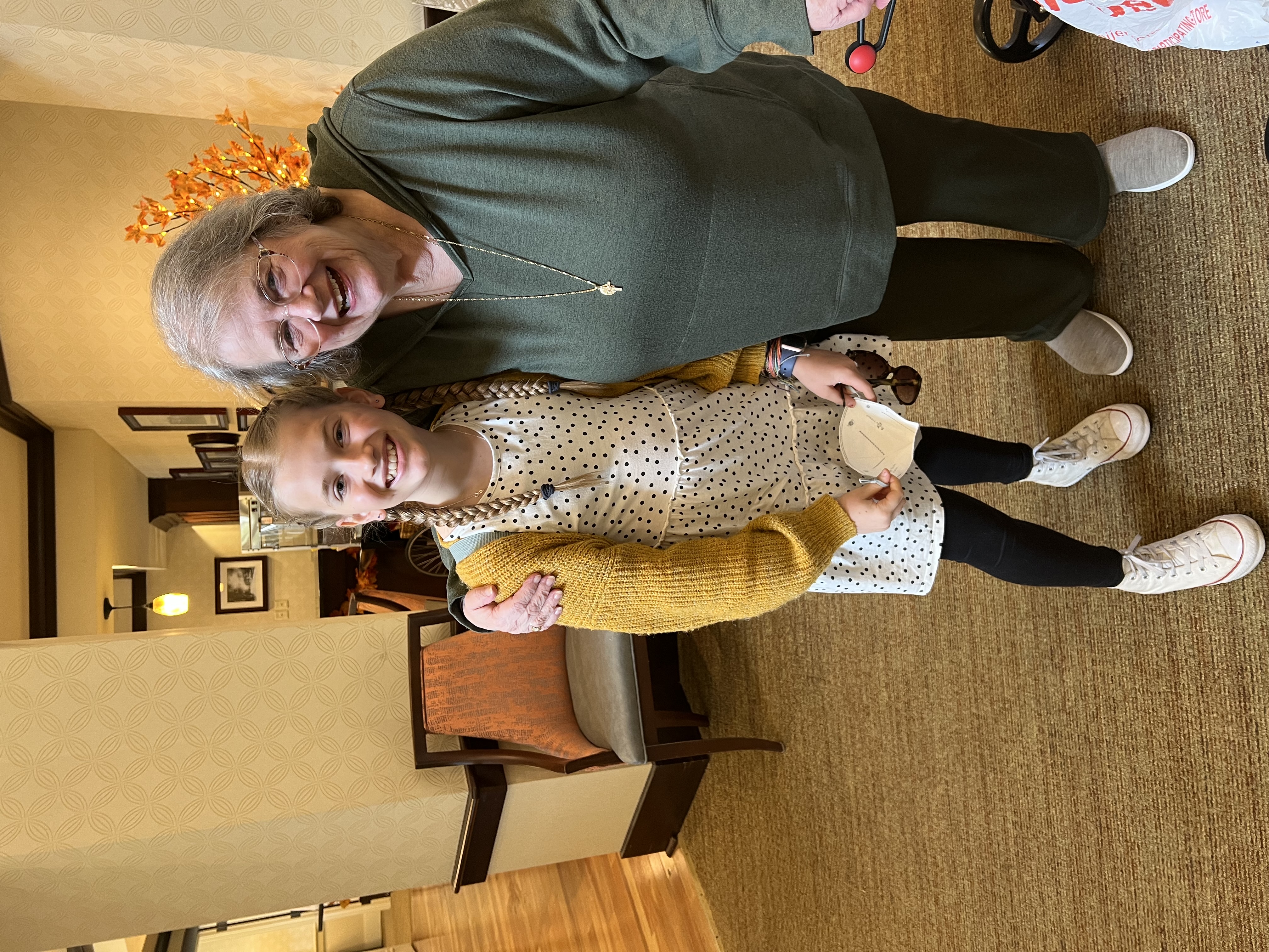 A smiling girl in a yellow cardigan with her arm around her grandmother in a residence hallway.