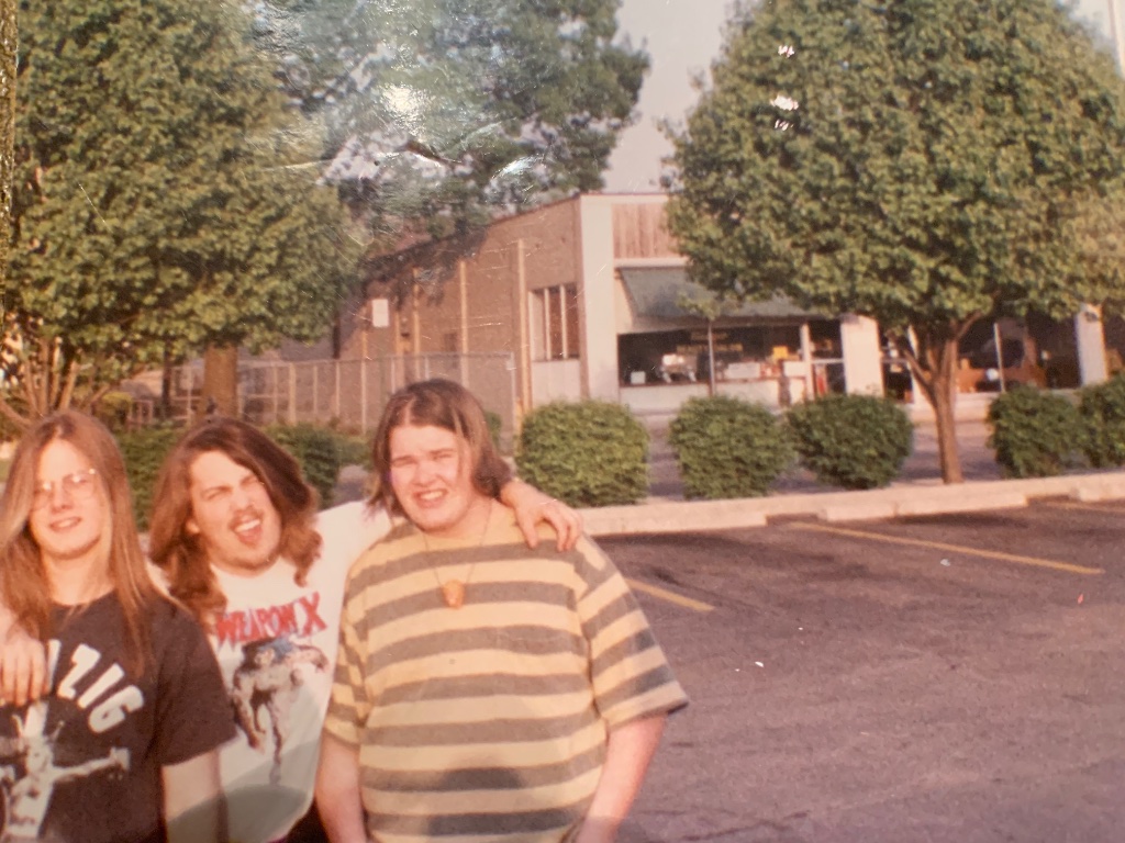 Three teenagers in band shirts in a parking lot, one with an arm around another's shoulder