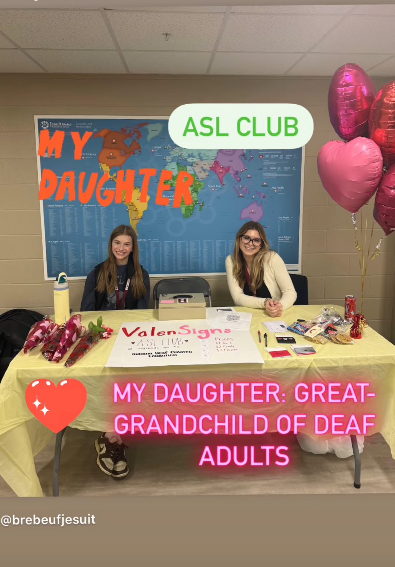 Two students seated at an ASL Club table with a hand-lettered ValenSigns sign, a world map, and heart-shaped balloons.