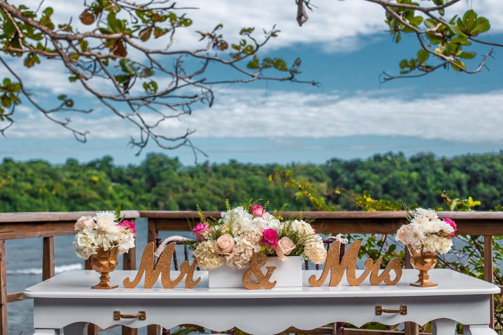 Mr & Mrs sweetheart table with gold glitter letters, pink and white floral arrangements, lush hillside and bay behind