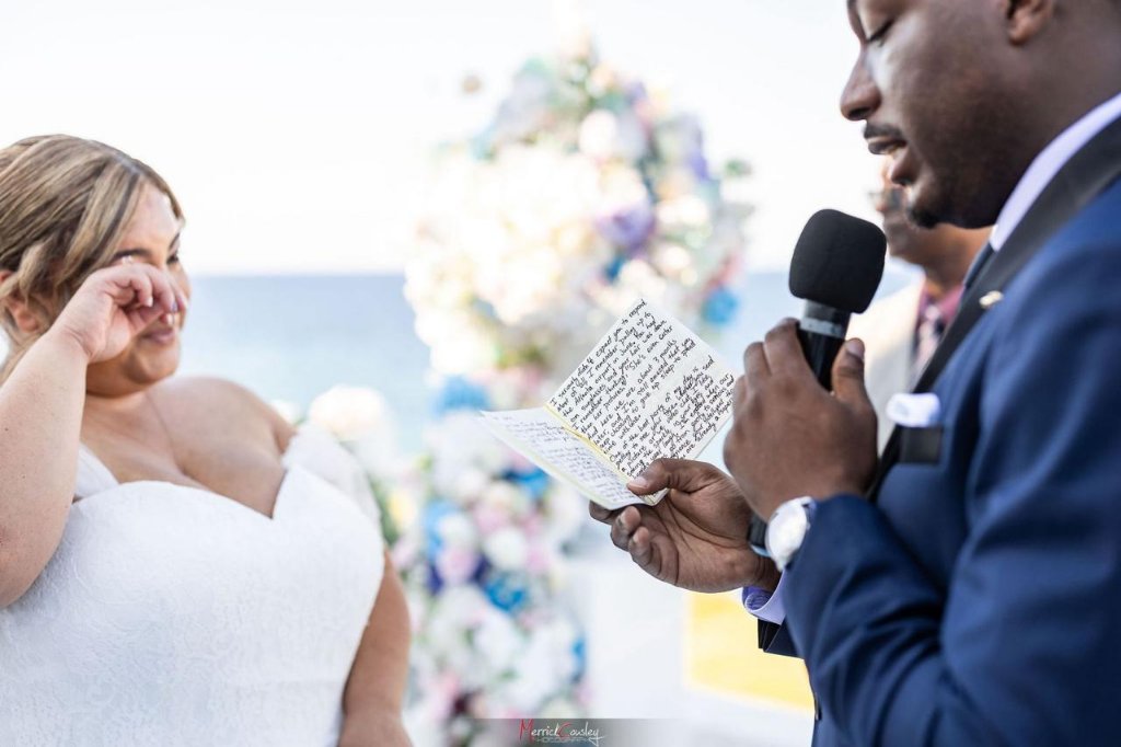 Groom reading handwritten vows at oceanfront ceremony, bride wiping happy tears