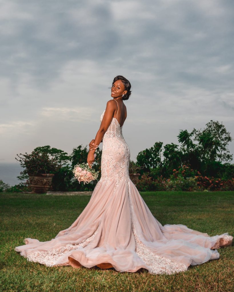 Bride looking over her shoulder, full blush train sprawling across the lawn, ocean glimpsed on the horizon