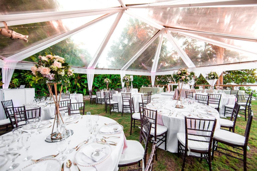 Interior of clear-roof reception marquee with round tables, white linens, dark chiavari chairs and tall gold centrepiece stands