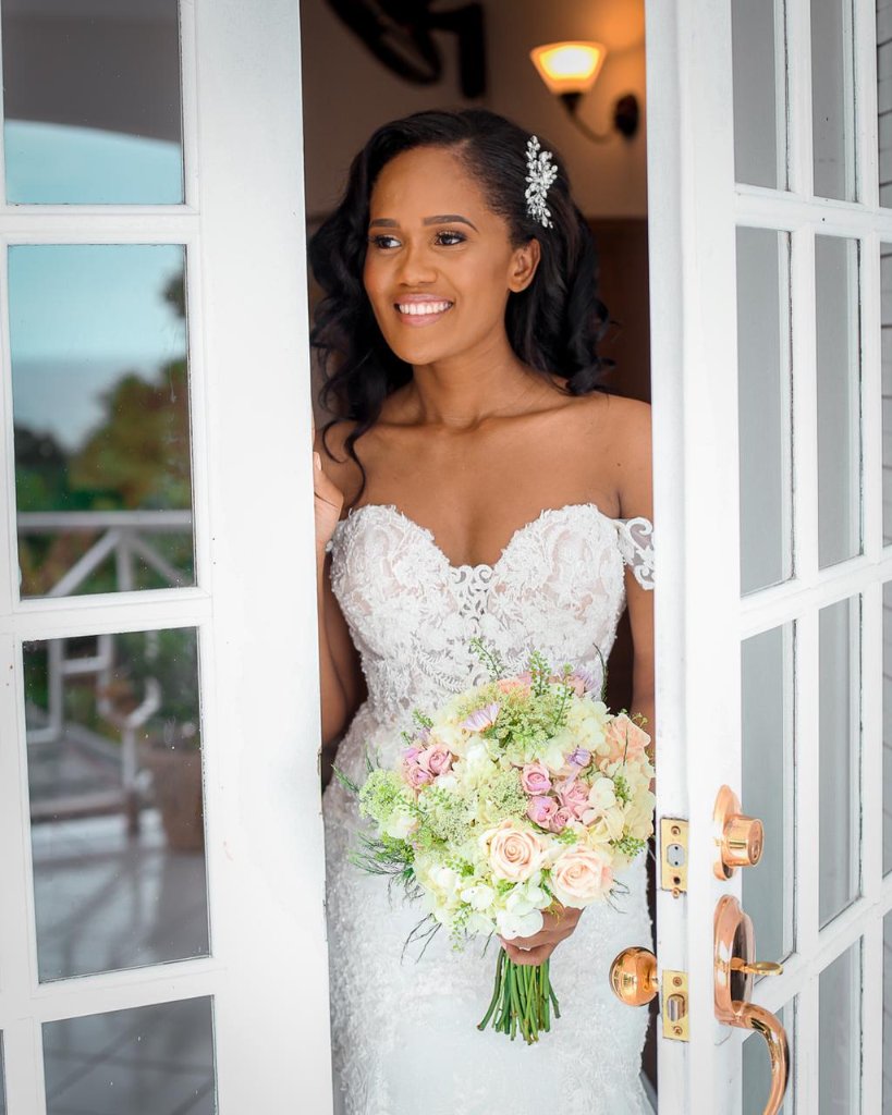 Bride smiling through open French doors in lace off-shoulder gown holding a blush bouquet