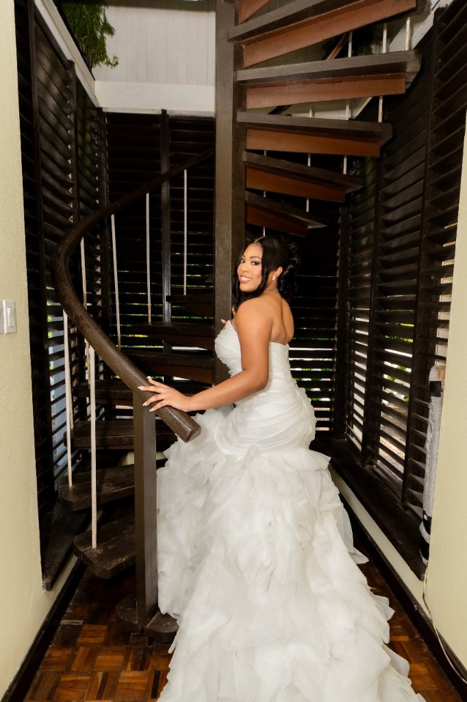 Bride in ruffled gown on dark wood spiral staircase, looking over her shoulder