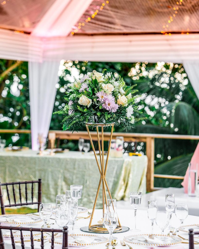 Gold geometric centrepiece stand topped with cream roses, lavender daisies and lush greenery under fairy lights inside the marquee