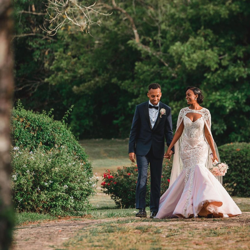 Couple walking hand-in-hand through estate garden grounds, both smiling, bride's blush train trailing, tropical blooms either side