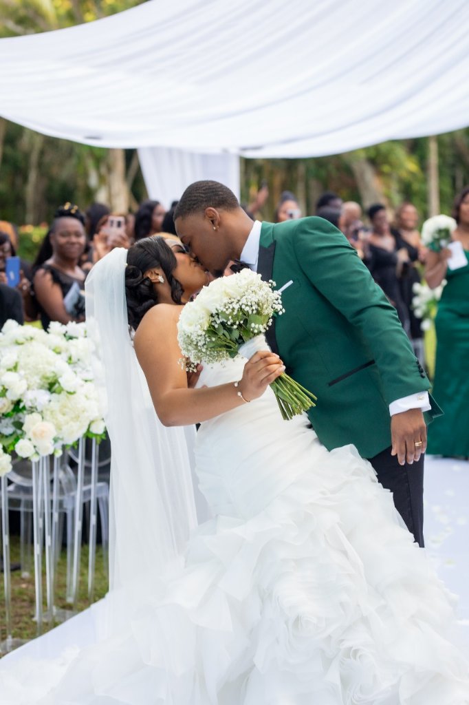 Couple sharing first kiss as recessional, crowd cheering behind them under white draping
