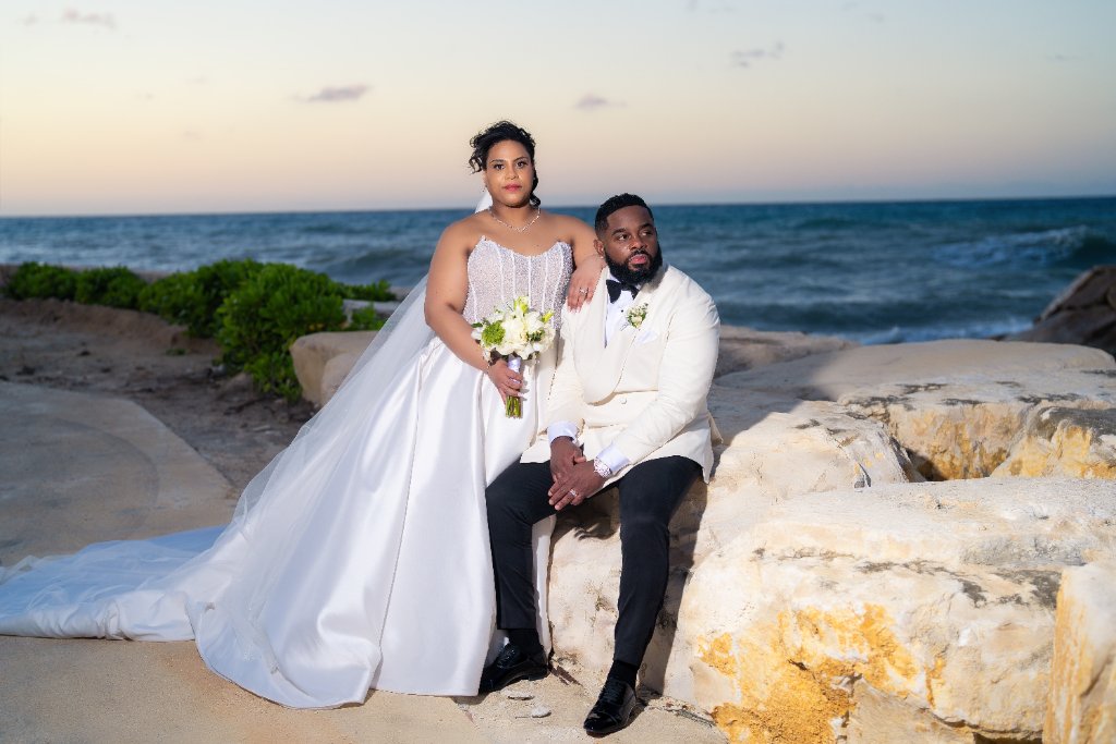 Couple seated on coastal limestone rocks at dusk, bride standing beside groom who sits casually, Caribbean Sea behind them