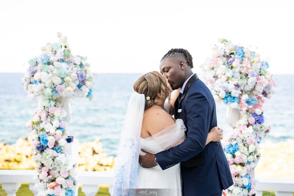 First kiss at oceanfront ceremony framed by pastel floral pillars at Trident Castle