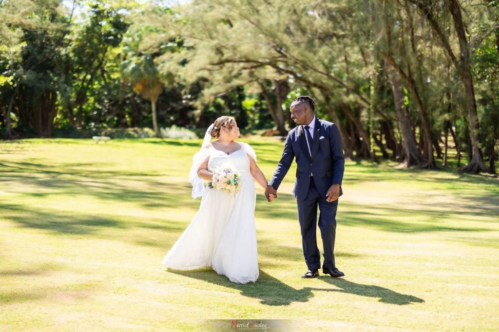 Couple walking hand-in-hand across sunlit lawn looking at each other, tree canopy above