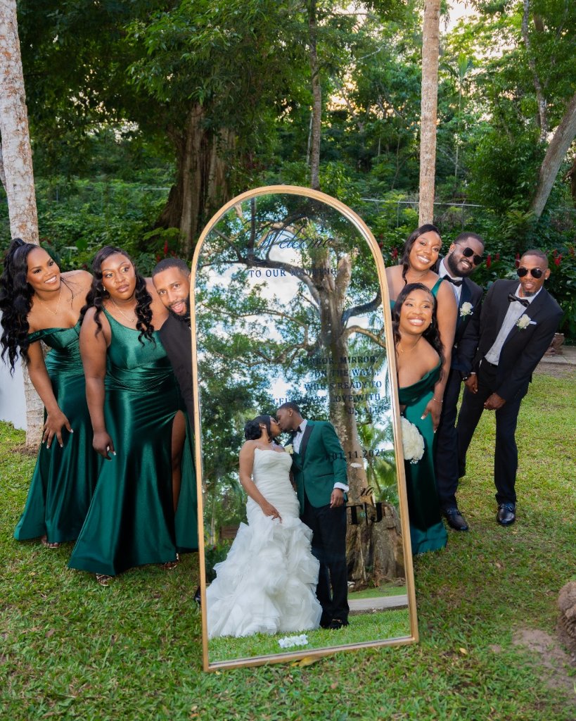 Wedding party posing around gold arch welcome mirror, couple kissing reflected inside, tropical garden