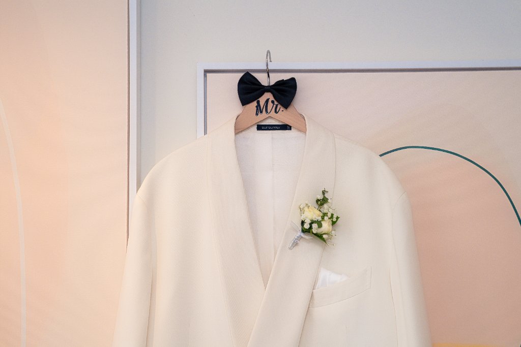 White Mr. suit jacket with boutonnière on custom wooden hanger with black bow tie against peach-toned hotel room wall