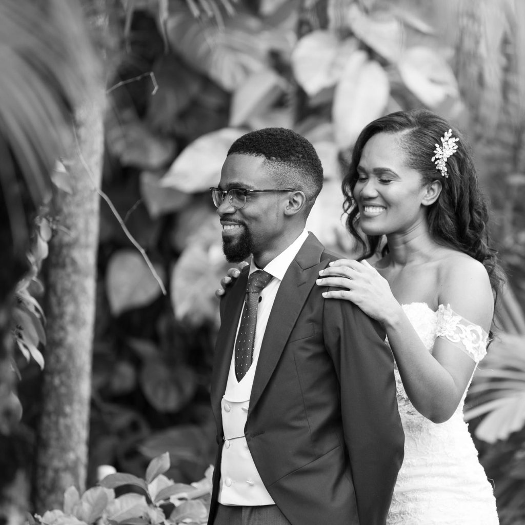 Black and white portrait of bride resting hand on groom's shoulder, both smiling, tropical foliage behind