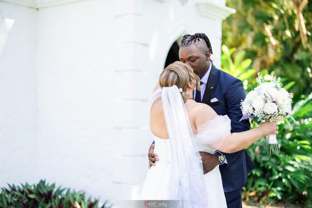 Groom kissing bride on the head outside white chapel, she holds bouquet
