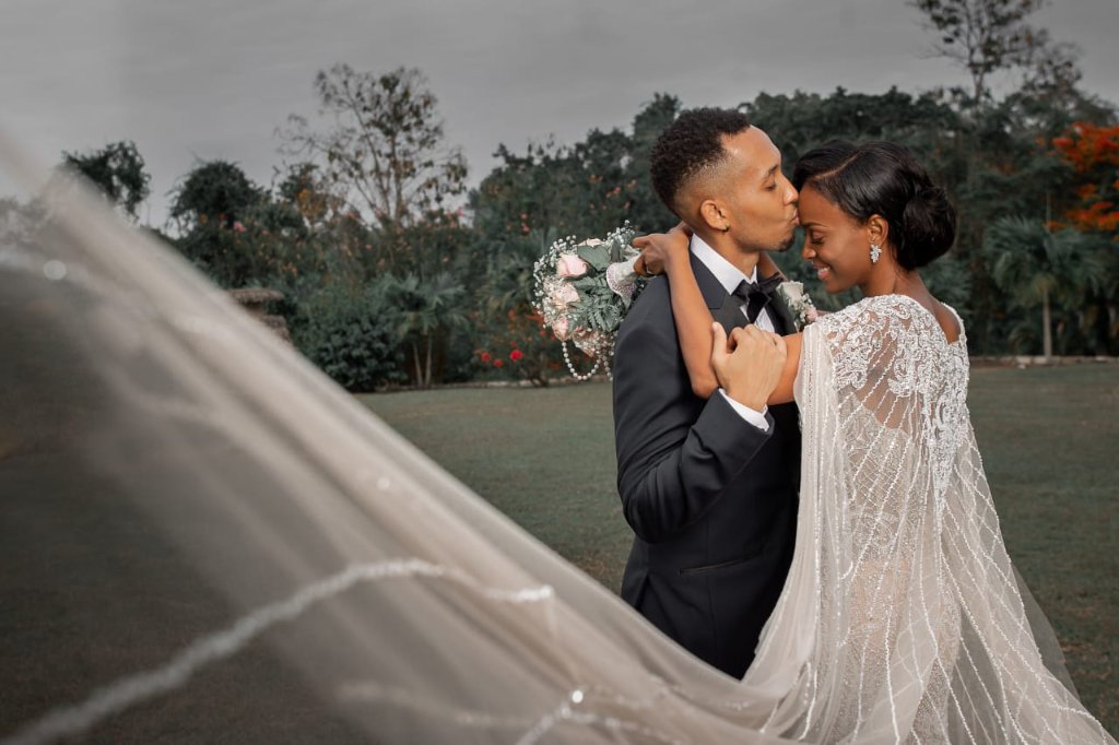 Groom kissing bride on the forehead, veil sweeping across the frame, lush garden and dramatic sky behind