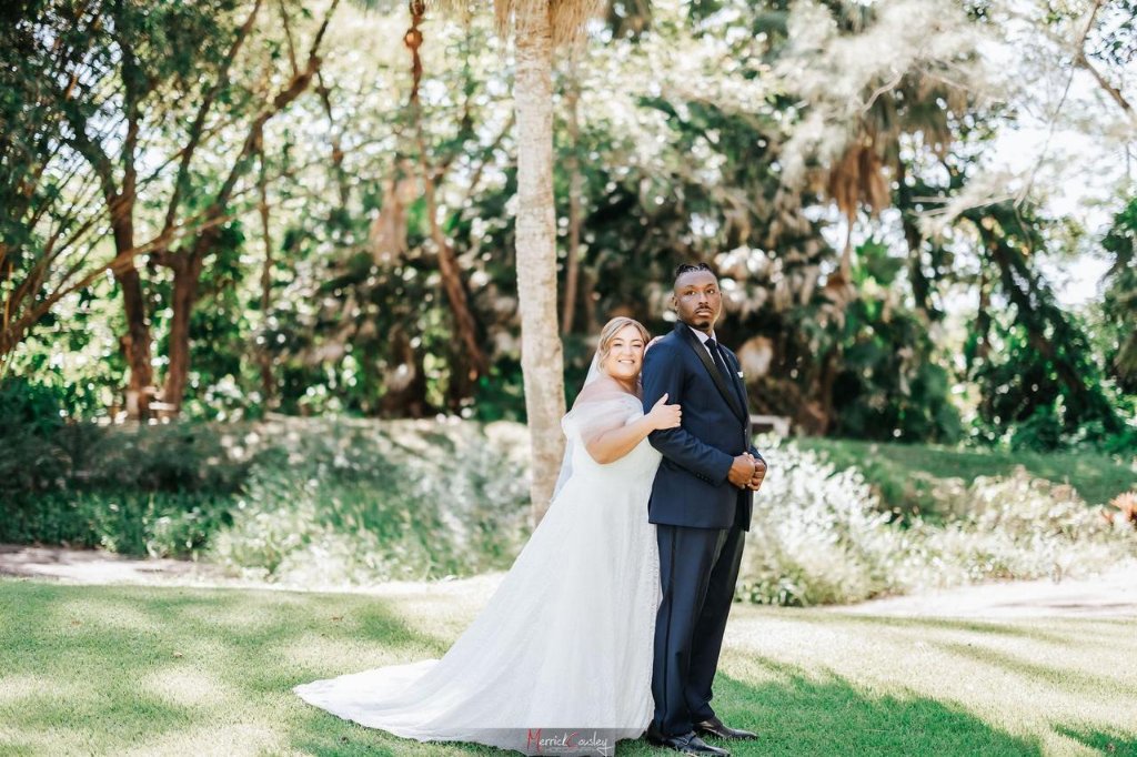 Couple back-to-back under palm tree in lush garden, bride smiling at camera