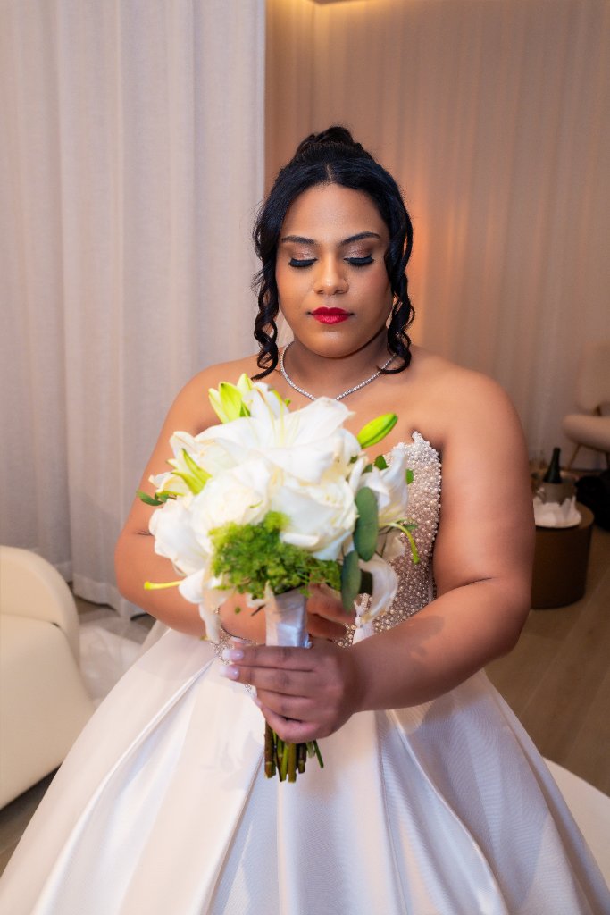 Bride in white ball gown holding white lily bouquet, eyes downcast, in warm-lit hotel suite