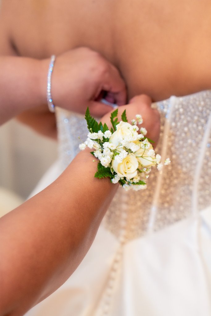 Close-up of bridesmaid's wrist corsage of cream roses and baby's breath as she fastens the back of bride's beaded gown